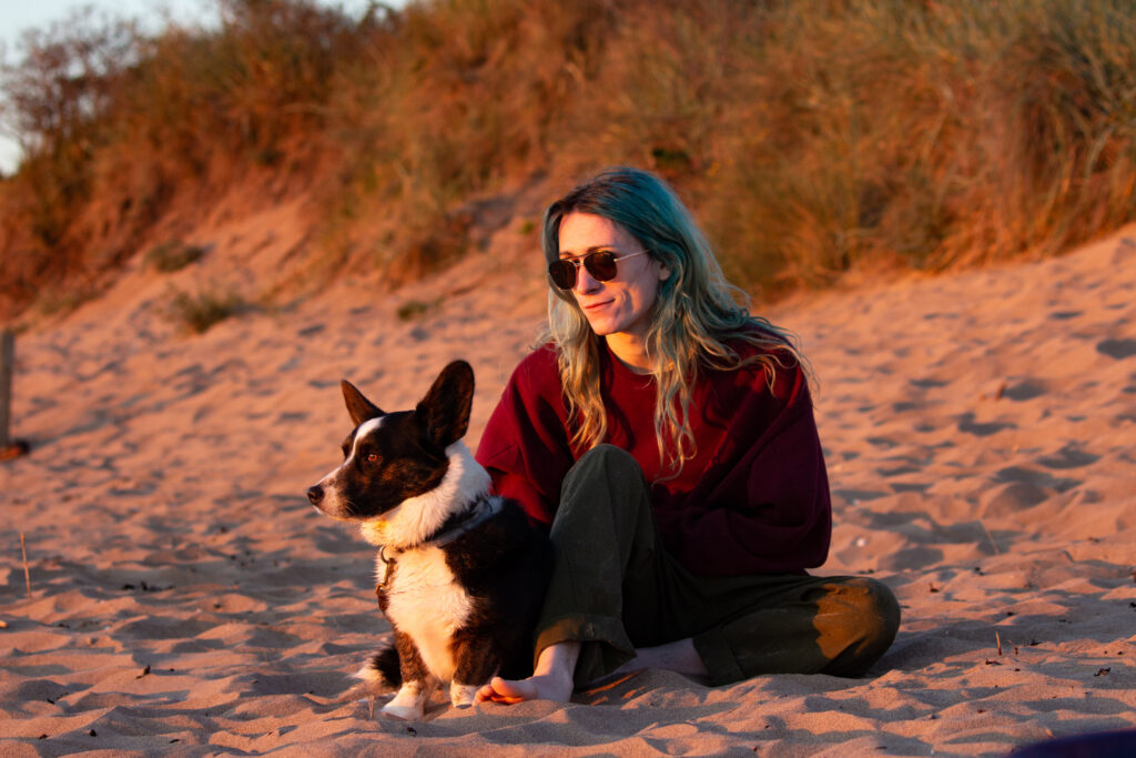 A person and a dog sitting on the beach, looking at a warm orange summer sunset.