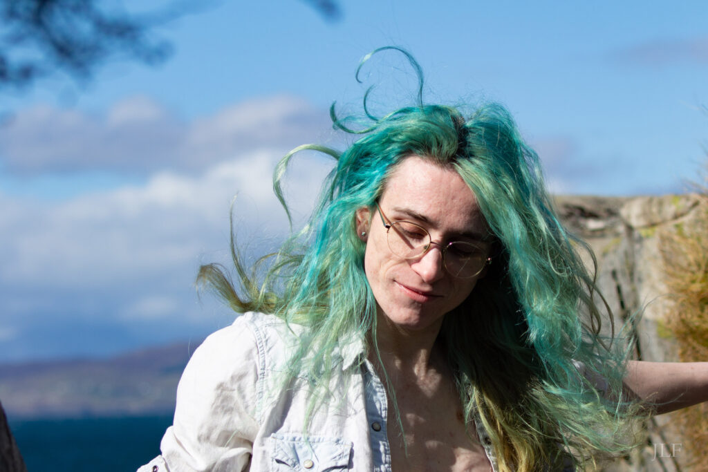 Close up portrait of a person in their late 20s at the beach. They are wearing glasses, long hair dyed teel blue and green, flowing in the wind