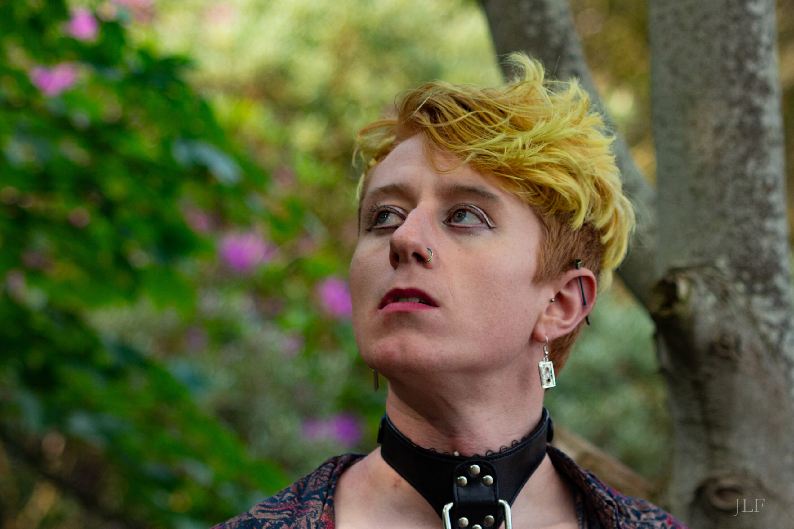 Portrait of Mitchell, in the woods. Yellow dyed short hair, make up, piercings, they are wearing a leather collar.