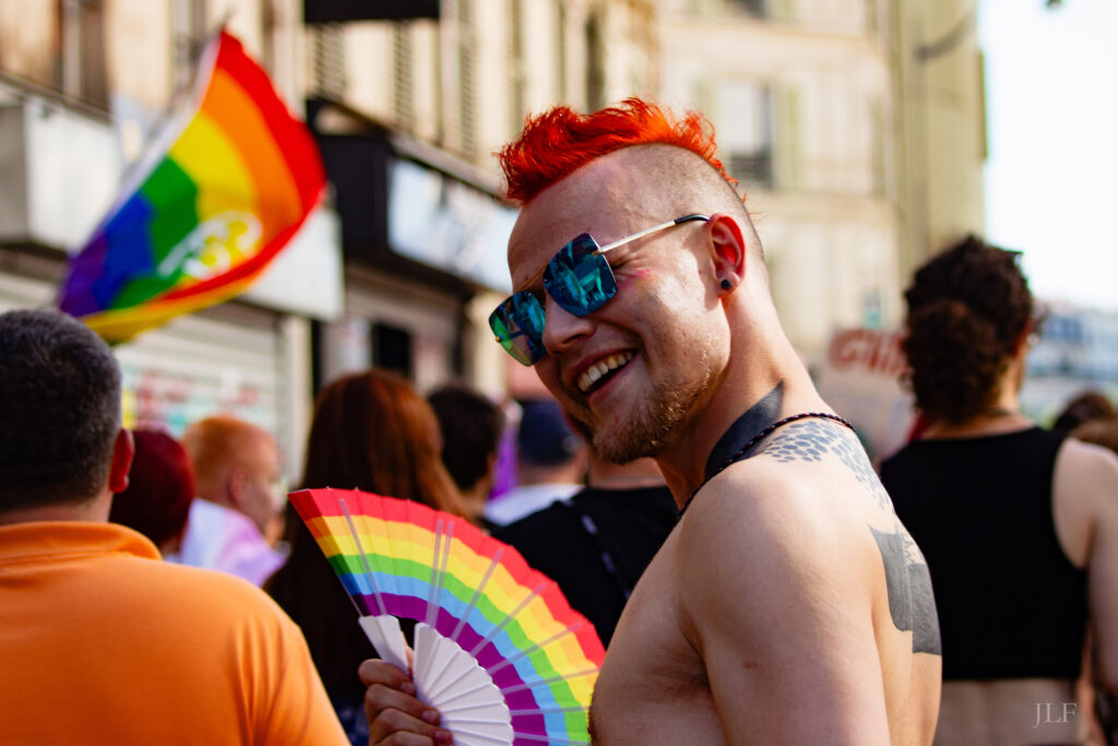 A person in the streets of Paris during the Pride march. red dyed hair, iroquois cut, wearing sunglasses and a rainbow coloured fan. 