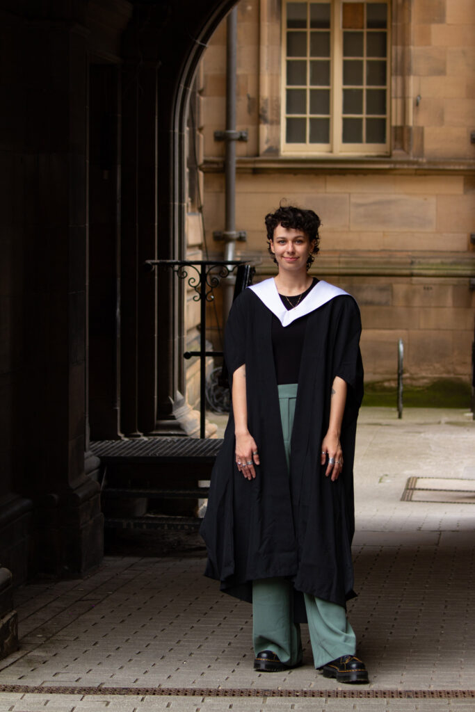 A person, short dark hair, standing under one of the stone arches at McEwan Hall. They're wearing their graduation gown over a black shirt and green trousers.