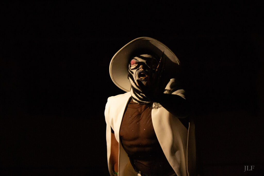 A person wearing a full face mask on stage during a drag show. Warm white side light gives very hard shadows on the performer's body.