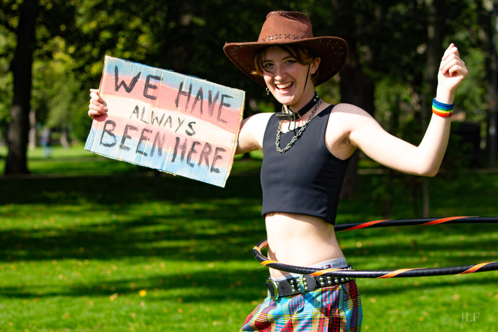 A person in the Meadows, Edinburgh, wearing a black crop top, rainbow wristband, holding a piece of cardboard with the trans flag colours and the words "We have always been here" painted in black.